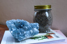 Load image into Gallery viewer, Large Blue Celestite cluster sitting atop a book with a jar of dried lavender in the background