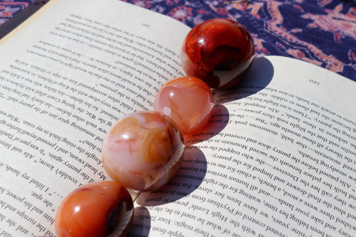 4 Carnelian eggs in various size variants lined up in the crease of an open book, colours varying from pinkish, orange citrus to deep reds and oranges
