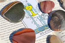 Load image into Gallery viewer, Polychrome Jasper Hearts in varying palm sizes, in shades of storm grey, green and warm oranges, sitting atop a book with a Tarot Card (Ace of Cups) in the centre 
