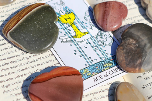 Polychrome Jasper Hearts in varying palm sizes, in shades of storm grey, green and warm oranges, sitting atop a book with a Tarot Card (Ace of Cups) in the centre 