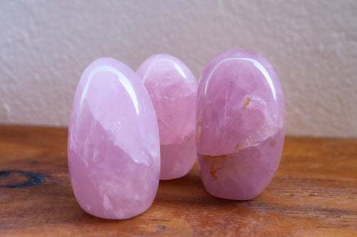 3 Rose Quartz boulders sitting together against a white wall on top a wooden bench. 