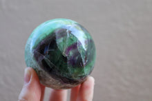 Load image into Gallery viewer, Very Unique, light coloured medium sized polished Fluorite sphere with Magenta and Green inclusions in the shape of an eye, held by fingertips

