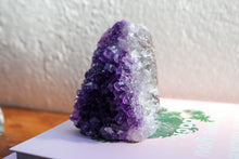Load image into Gallery viewer, Medium sized triangular amethyst cluster sitting atop two books, combined dark purple to lilac, facing away from light source
