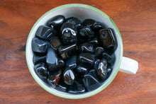 Load image into Gallery viewer, A large pile of Shiny Black Agate tumbles showing red and white inclusions sitting in a large soup dish
